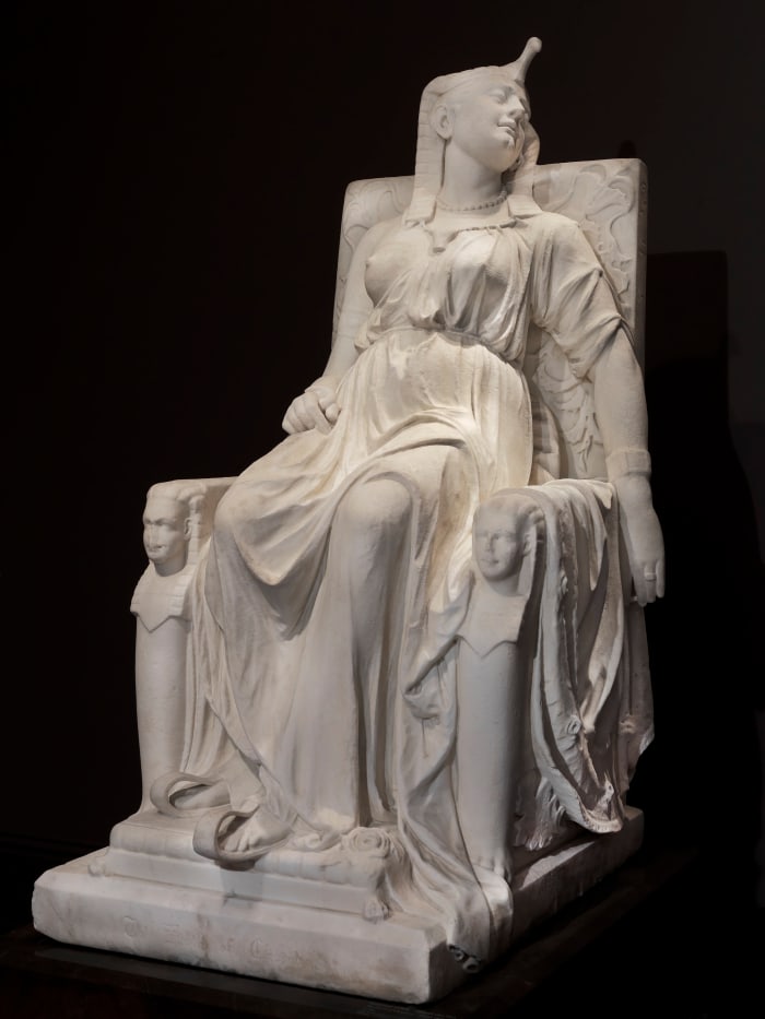 “The Death of Cleopatra,” carved 1876, marble, 63” x 31-1/4” x 46”, now in the Smithsonian American Art Museum. The legendary queen of Egypt is often best known for her dramatic suicide, allegedly from the fatal bite of a poisonous snake. Here, Lewis portrayed Cleopatra in the moment after her death, wearing her royal attire, in majestic repose on a throne. The identical sphinx heads flanking the throne represent the twins she bore with Roman general Marc Antony.