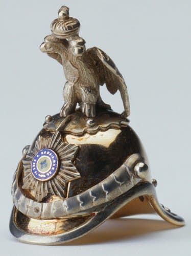 Fabergé made numerous objects with military associations, often as presentation pieces. Helmets were produced as drinking vessels, although this miniature version made before 1896 by Erik August Kollin is far too small to have been intended as a functional object. It is a helmet of the Imperial Chevalier Guard and is enameled on the front with the star of the Order of St Andrew. The helmet is surmounted by the imperial eagle; 1” x 1” x 1/2”.
