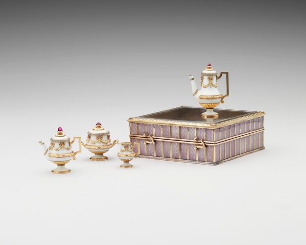 This gold, crystal, enamel and ruby tea service was made by Carl Fabergé, 1896-1908; 1” x 2-1/2” x 2-1/2”. Comprising a teapot, hot water pot, sugar bowl and milk jug, this tea set is of gold enameled in a pale opaque bluish-white, but there is no engine turning on the metal. This deliberately plain enameling creates the impression that the tea set is made of porcelain. The lids are each surmounted by a finial in the form of a cabochon ruby. The quality of the gemstones and the rich application of four colored gold decoration may indicate that this was an imperial commission. The box is of silver-gilt, applied with colored gold and with guilloché enamel in mauve, the favorite color of Tsarina Alexandra Feodorovna, which this belonged to, and the lid is set with a panel of crystal.