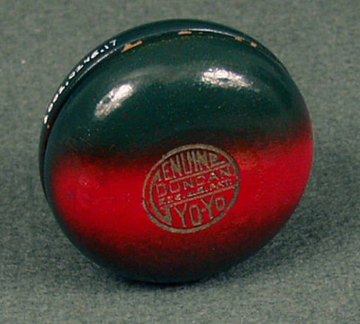 A wooden yo-yo made by the Duncan Toys Company, 1930s. It has a green design with a broad red stripe. The seal reads “Genuine Duncan Yo-Yo, Reg. US Pat.” This is an early version of the Duncan Genuine Yo-Yo, produced soon after Donald Duncan bought the trademark term “yo-yo” from inventor Pedro Flores, and the seal is reminiscent of the one used by Flores.