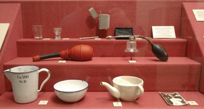 This display case in the Living History Museum showcases some of the items nurses needed when caring for private duty patients. The minim cup and eye cup at upper left allowed for the precise administration of medications, while the metal case protected glass syringes and their needles. On the center level, rubber douches and atomizers alike provided easy irrigation of wounds or sore throats. And on the bottom, enamel ware jugs and bowls were useful in preparing the kinds of soft foods used in the ceramic “pap boat.”