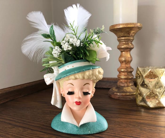 Lucille Ball lady head vase, Napco, stamped C3959 ©NAPCO 1959, foil label is partially missing, bristle-fiber eyelashes, aqua green coat with white upturned, glazed collar and a wide-brimmed hat with polk-a-dot bow, 5-1/2”. A crown of faux roses, greens, and feathers created by the seller is removable; $99.97.