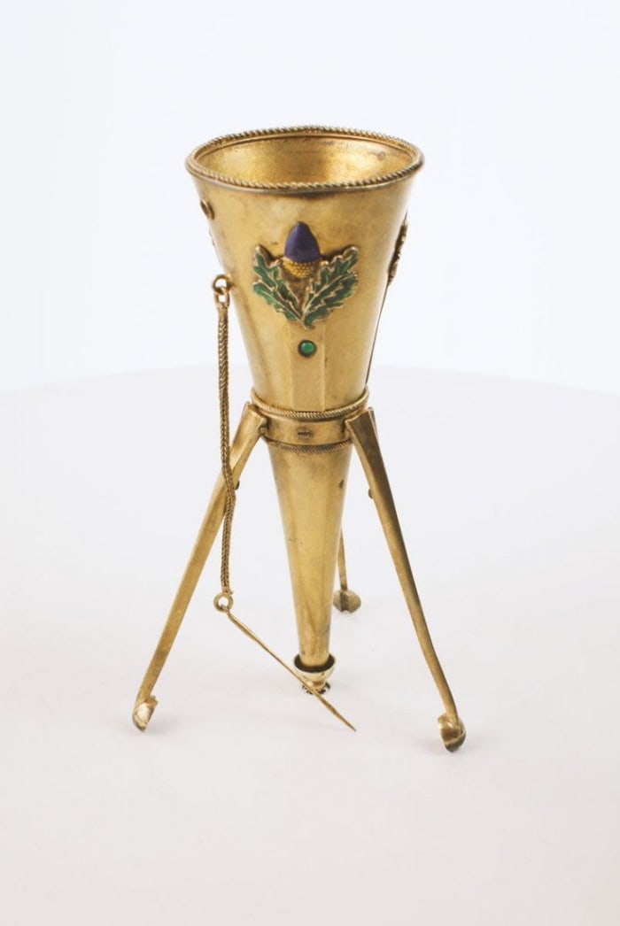Funnel-shaped tripod holder, circa 1880, silver with a gold wash, raised purple enamel flowers with green leaves on three sides, floral pin attached at the neck, legs collapse into a small spring-loaded retaining cup at the tip when carried in the hand, 4-1/2” h; $2,600.