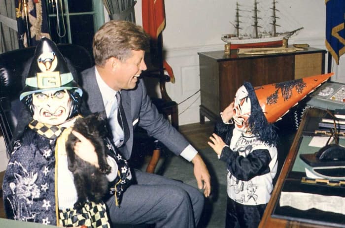 President John F. Kennedy laughs as his children, Caroline Kennedy (left, holding a cat) and John F. Kennedy, Jr., model their Halloween costumes in the Oval Office.