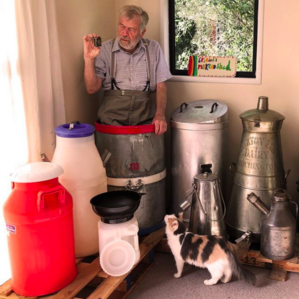 Ian Spellerberg is standing in his largest milk can (25 gallons) from New Zealand and is holding the smallest milk can in the world (1/8th of a pint) from England. His adopted cat Florence (found as a kitten on a country road) watches with interest. Also shown are three modern plastic milk cans on the left. On the right there are two unusual cans with spouts.
