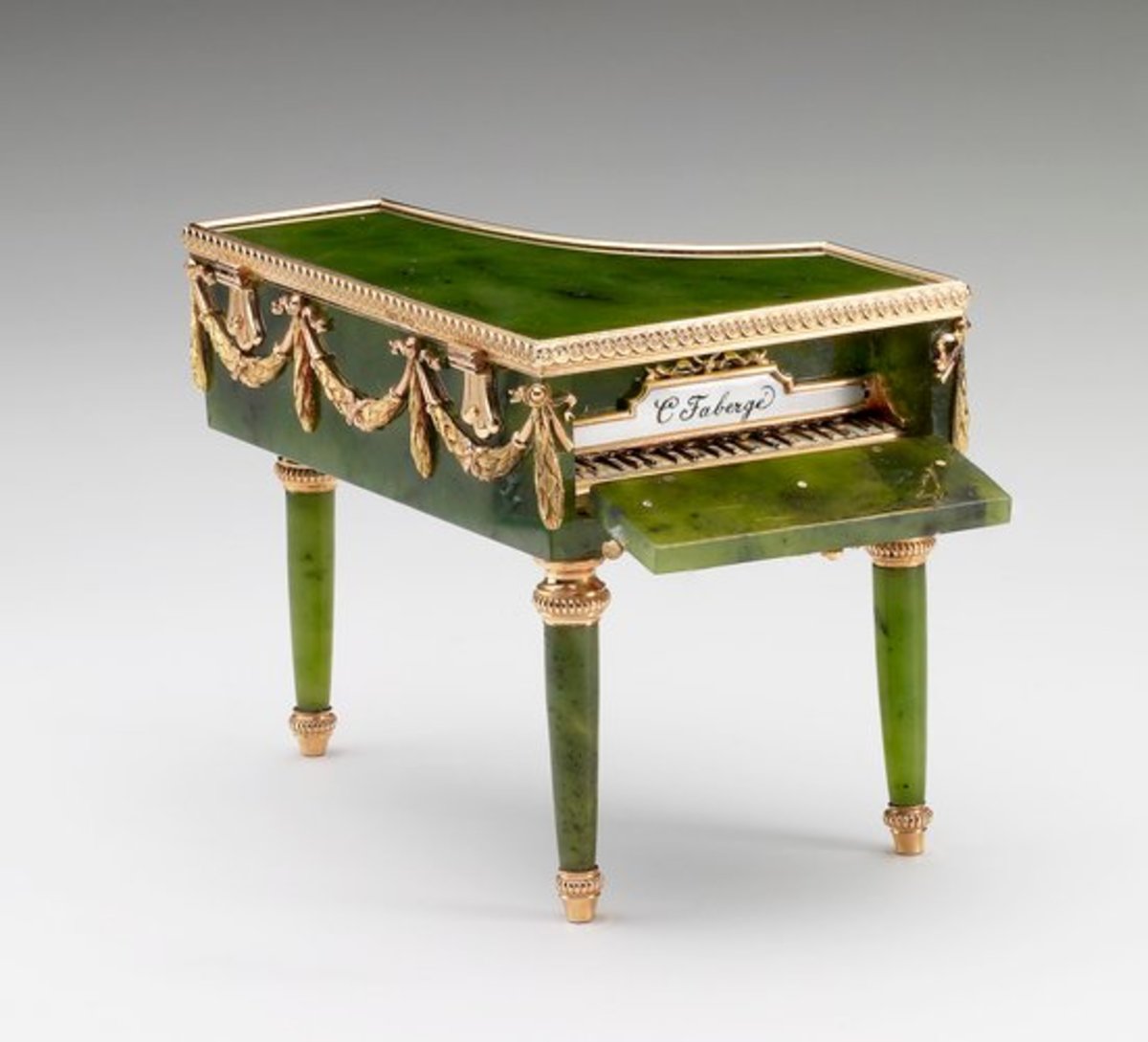 This miniature grand piano in Louis XVI style was made by Mikhail Perkhin. It’s made of nephrite with mounts of red and green gold, and the keyboard is opaque black and white enamel. Foliate swag mounts are around the edge and the top is hinged; 2” x 2.8” x 2”. Judging by her acquisitions, Queen Mary was particularly fond of Fabergé’s miniature objets de fantasie, which include several examples of miniature furniture in the form of bonbonnières. These objects afforded the craftsman the opportunity to demonstrate skill in applying specialist techniques to replicate the real materials of the full-scale object. This miniature piano of Siberian nephrite is carved and polished to resemble ebonized wood. The lid opens for use as a bonbonnière and the front drops down to reveal the keyboard in gold and enamel, inscribed “C. Fabergé.” The piano belonged originally to Tsarina Alexandra Feodorovna and is also seen in one of the display cases in photographs of the exhibition of Fabergé held in St. Petersburg in 1902. Queen Mary acquired it sometime between 1922-1931.