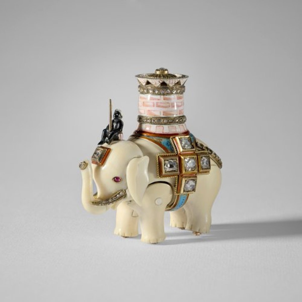 Mikhail Perkhin made this ivory elephant automaton in 1892; 2-1/2” x 2” x 1-1/2”. The elephant has separate, jointed body legs and head and is ridden by an enamel man sitting on its head and carrying a pink and white enamel castle with two rose-cut diamond set bands and a pierced upper rim; the top set with a single diamond in gold. This automaton was made as the “surprise” for the Diamond Trellis Egg, made by Carl Fabergé for Tsar Alexander III. The Tsar presented the egg to his wife, Tsarina Maria Feodorovna, for Easter 1892. The elephant is wound with a watch key through a hole hidden underneath the diamond cross on one side of the elephant. It walks on ratcheted wheels and lifts its head up and down. You can learn more and watch a short video about this piece here.