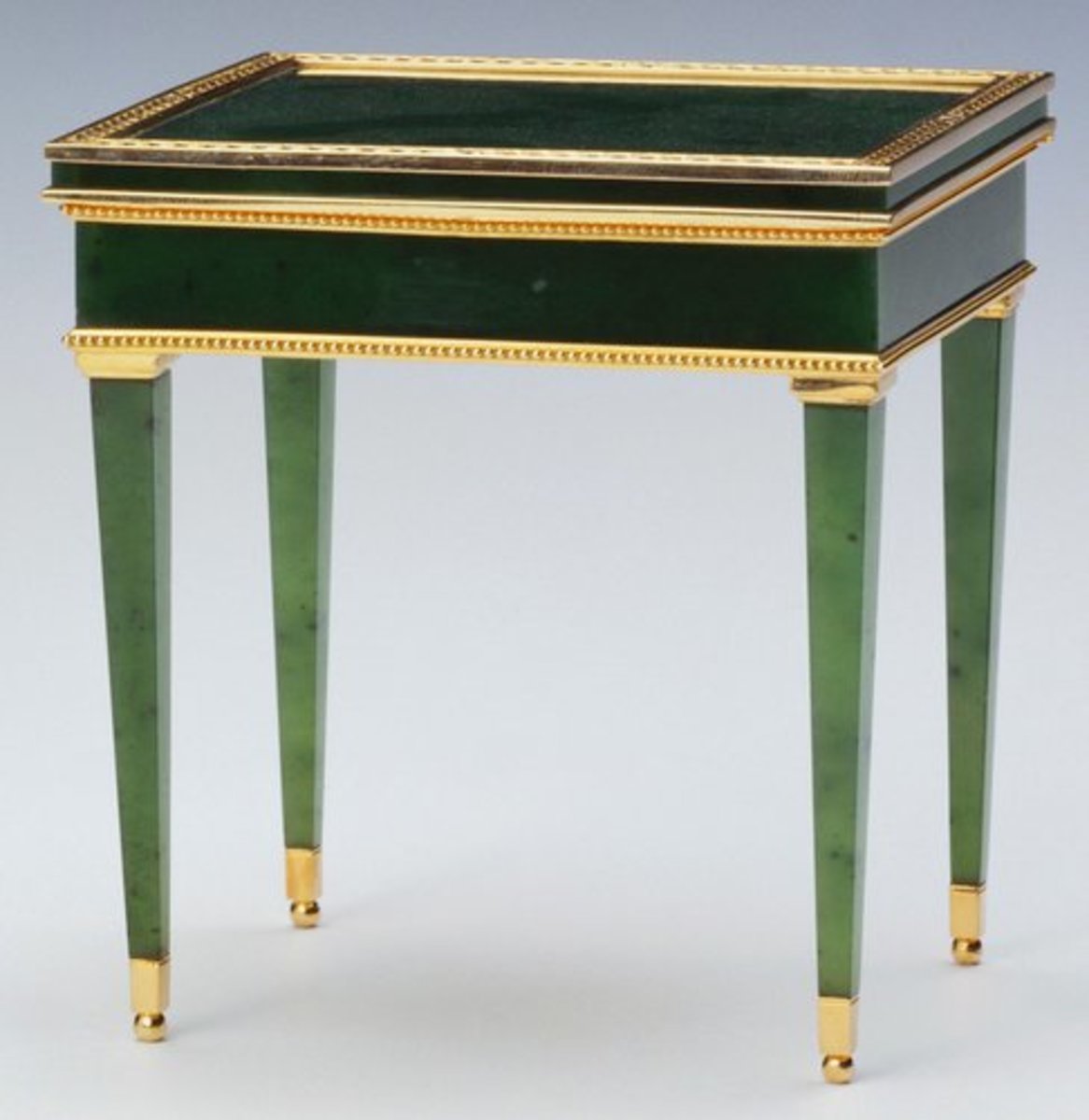 Rectangular miniature Louis XVI-style table by workmaster Mikhail Perkhin, 1896-1903, is made of nephrite and mounted with red gold borders and has tapering legs with gold feet and a chased lid with leaves around edge and is hinged; 3” x 2.7” x 2”. Fabergé produced a number of bibelots in the form of miniature furniture. Some, such as this table, were intended to be used as bonbonnières, fancy boxes for bonbons. This was a Christmas present for Queen Mary by Lord Revelstoke in 1921.