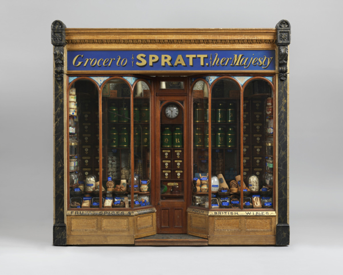 Model of a grocery store called “Grocer to SPRATT Her Majesty,” with a glazed door and windows and a display of miniature baskets, jars and bottles of food. Inside is a wooden counter and at the back of the store are wooden drawers and metal jars that contain spices and groceries. The whole model is in a glass case; 28-1/2” x 32” x 24-1/2”.