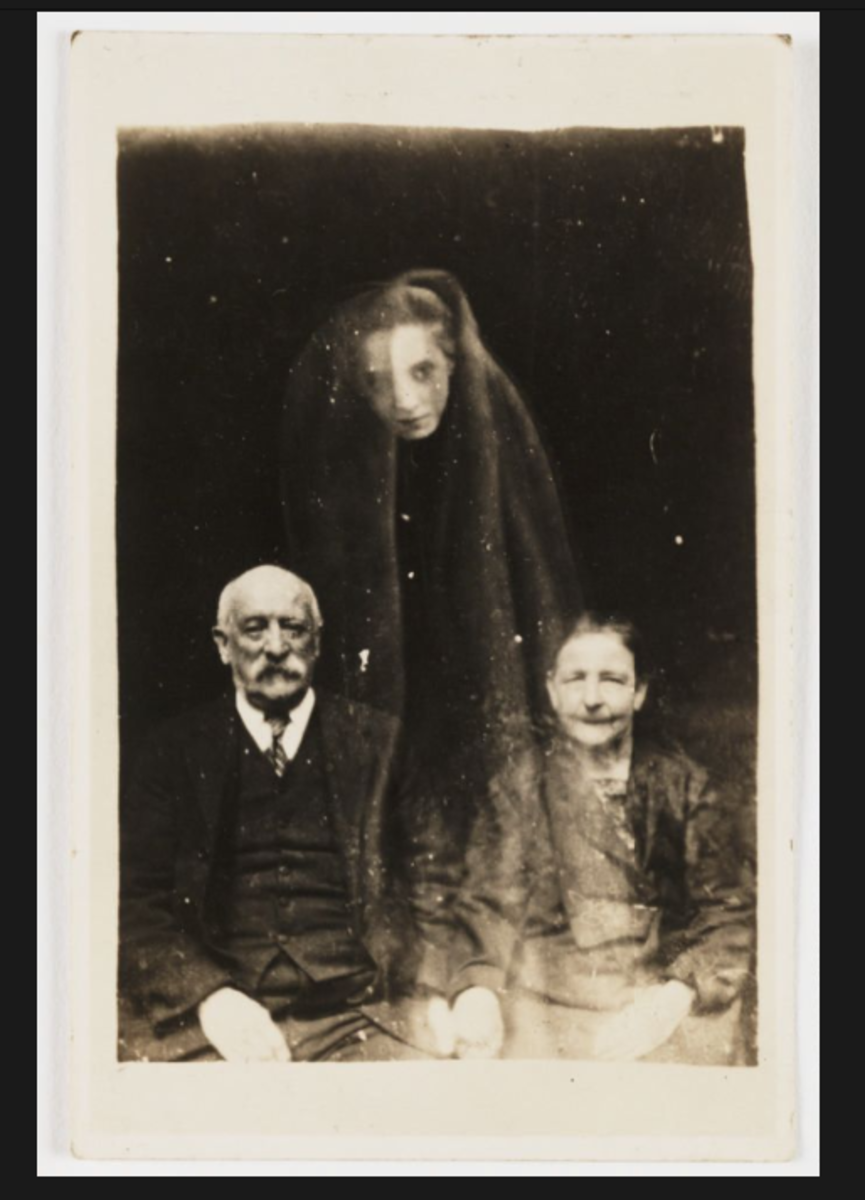 A portrait photograph of a seated elderly couple, possibly taken by William Hope in about 1920. A young woman's face appears as if floating above them, draped in a cloak.