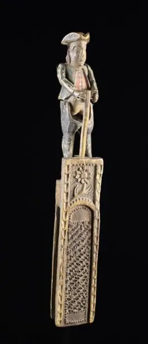 A rare 18th century carved and polychrome automata nutmeg grater, possibly a reference to the East Indian spice wars. The pierced sheet iron rasp set in a carved frame decorated with a daisy and surmounted by an articulated figure of man wearing a tricorn hat pounding a pole, with his trousers dropping to expose his backside when bending, 10-1/2” h; $7,760.