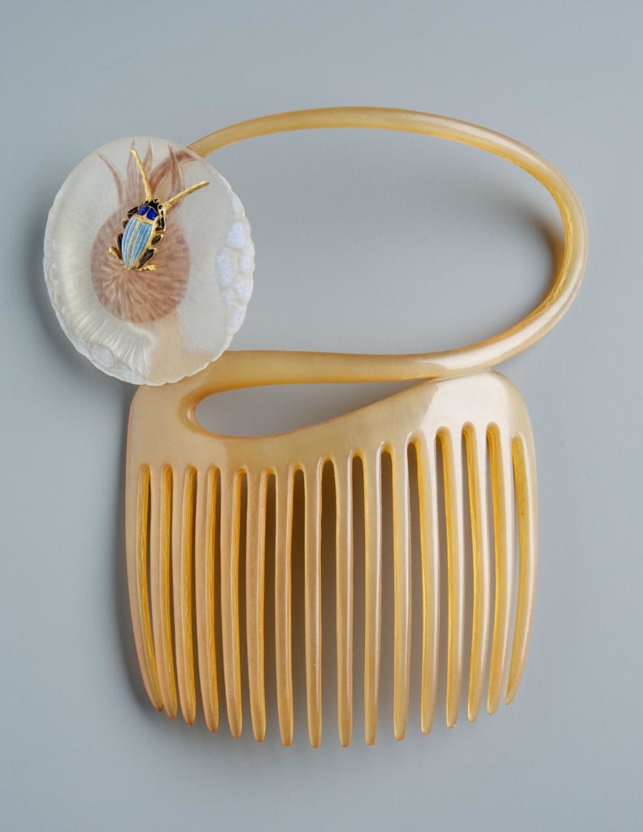 This comb, circa 1898-1899, is sawed of a light, transparent horn. Its curved top is continued in a tendril that recoils in an upturned S-shape. The tendril ends in a round, puffy blossom of a lion's tooth made of matte-etched glass. There is an enameled, naturalistic insect sitting in the center of the flower: a golden bug with two long feelers, a blue chitin cover and black legs. The rim of the petal is of irregular thickness, being blue opaque at the wider places. This comb is also a typical example of Japanese influence of Lalique's work; 4.3" h, 3.9" w.