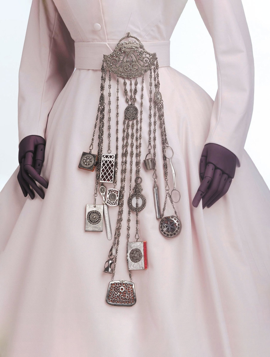 An elaborate cut-steel and ivory chatelaine, circa 1863-65, shown as it would have been worn attached to a dress. Tools include scissors, scent bottle, case, case top, miniature bucket, needle case, possibly a cigar cutter, paper knife or letter opener, writing tablet case, notebook and magnifying glass.